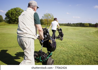 Active senior golf couple pushing golf trolley with bag on fairway on beautiful golf course with blue sky in background. - Powered by Shutterstock