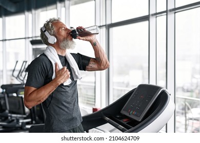 Active Senior In Grey T-shirt With White Towel On Shoulders, Drinking Water From Bottle, Standing On Treadmill. Aged Man With Headphones Staying Hydrated After Sport Session.