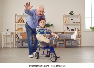 Active Senior Man Feels Young Again When Plays Carefree Childish Games With Grandson. Happy Little Kid Having Fun, Riding Tricycle And Fooling Around While Enjoying Free Time With Grandpa At Home