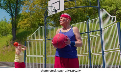 Active Senior Man Grandfather 80 Years Old Posing With Ball, Looking At Camera Outdoors On Playground Court. Healthy Grandmother Playing Basketball In Background. Sport Motivation For Elderly People
