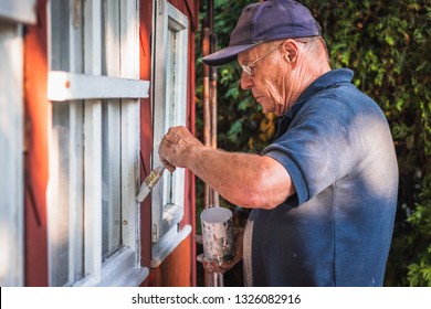 Active Senior Man Painting Window Frame Of Wooden Cabin. House Improvemnet. Old Craftsperson Repairing House Exterior. 