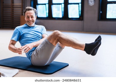 An active senior man is smiling while doing a sit up on a mat. He is wearing a blue shirt and gray shorts
