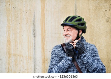 Active senior man standing outdoors against a concrete wall, putting on bicycle helmet.