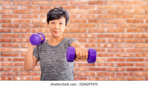 Active Senior Older Asian Attractive Elder Woman Training, Exercising, Workout At A Gym Club With Lifting Weights Dumbbells Over Old Red Brick Background. Older Senior Adults Exercise Concept Ideas.