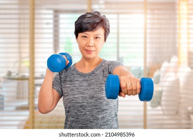 Active Senior Older Asian Attractive Elder Woman Training, Exercising, Workout At Home With Lifting Weights Dumbbells And A Bedroom On A Background. Older Senior Adults Exercise At Home Concept Ideas.