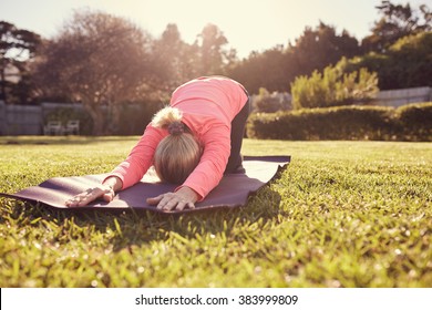 Active Senior Woman Bent Over In Child's Pose On A Yoga Mat, Spread Out On Fresh Green Grass In Her Garden On A Summer Morning, With Gentle Sunlight