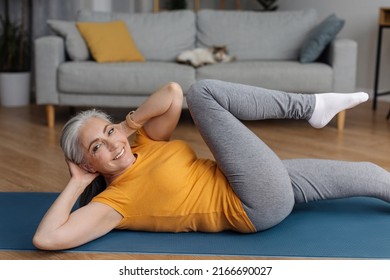 Active Senior Woman Doing Abs Exercises On Yoga Mat At Home And Smiling At Camera. Sporty Mature Lady Working Out In Living Room Interior. Strength Training Concept