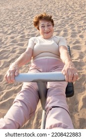 Active Senior Woman Doing Sports Exercises Outdoors. Pensioner Does Stretching On Beach Using Exercise Equipment. Concept Of Physical Activity In Old Age, Health, Healthy Lifestyle, Fitness. Autumn.