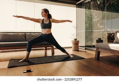 Active senior woman doing the warrior pose during a session of yoga. Mature woman doing a balance exercise on a yoga mat. Sporty woman practicing a healthy workout routine at home. - Powered by Shutterstock