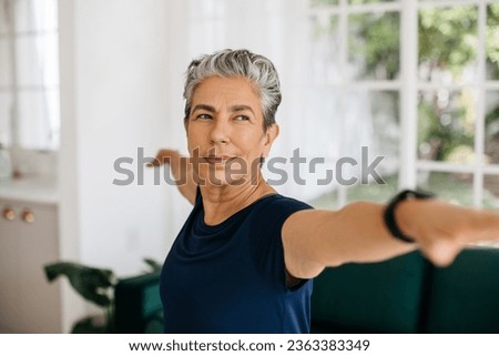 Active senior woman doing the warrior pose at home, she stretches her arms with a confident look on her face. Mature woman practicing yoga to improve her strength, balance and stability.