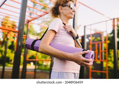 Active Senior Woman With Fitness Mat And Water Bottle In Hands Walking On Workout Outdoor Gym. Happy Mature Lady Strolling In Sports Ground Going At Outdoors Yoga Class. Active Lifestyle Concept.