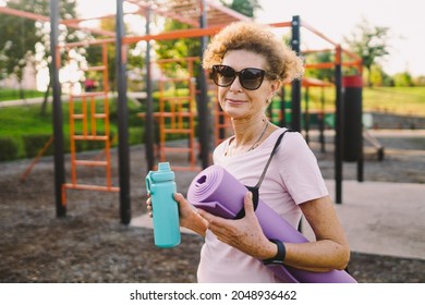 Active Senior Woman With Fitness Mat And Water Bottle In Hands Walking On Workout Outdoor Gym. Happy Mature Lady Strolling In Sports Ground Going At Outdoors Yoga Class. Active Lifestyle Concept.