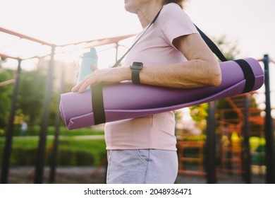 Active Senior Woman With Fitness Mat And Water Bottle In Hands Walking On Workout Outdoor Gym. Happy Mature Lady Strolling In Sports Ground Going At Outdoors Yoga Class. Active Lifestyle Concept.