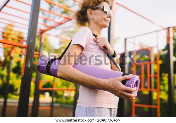 Active senior woman with fitness mat and water\
bottle in hands walking on workout outdoor gym. Happy mature lady\
strolling in sports ground going at outdoors yoga class. Active\
lifestyle concept.