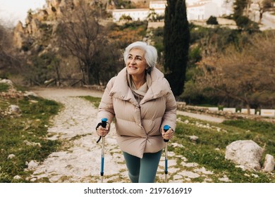 Active senior woman hiking with trekking poles outdoors. Adventurous elderly woman smiling while walking up a hilly trail. Mature woman enjoying recreational activities after retirement.