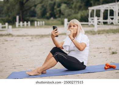 Active senior woman hydrating and using her smartphone while taking a break from her outdoor exercise routine at the beach. - Powered by Shutterstock