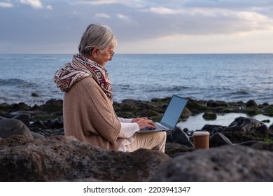 Active Senior Woman Sitting On The Rocks Beach Using Laptop. Elderly Caucasian Female In Online Remote Work Enjoying Sunset At Sea, Horizon Over Water