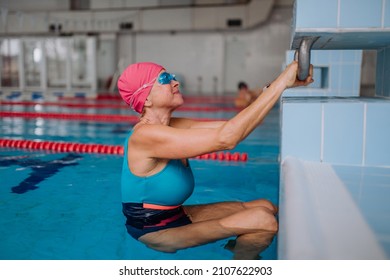 Active Senior Woman Swimmer Holding Onto Starting Block Preparing To Swim In Indoors Swimming Pool.