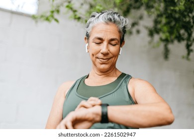 Active senior woman using a smartwatch to set a timer before going for a morning run. Woman with grey hair starting her outdoor workout routine, using technology to track her fitness progress.