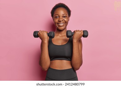 Active slim woman dressed in black cropped top and leggings lifts dumbbells being in good physical shape smiles pleasantly isolated over pink background doing biceps curl. Healthy lifestyle concept