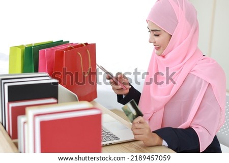 Active smart asian woman in blue muslim suit and pink shaft sitting and holding credit card while using mobile phone for payment or online purchasing. Startup small business girl work at home.