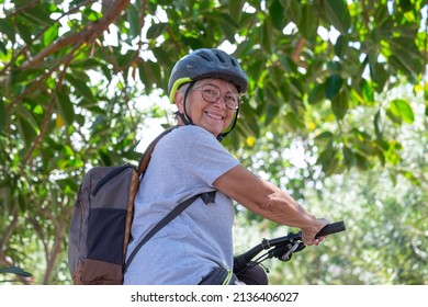 Active Smiling Senior Woman Cycling With Her Electro Bike In A Green Park Wearing Helmet Looking Back. Mature Attractive Woman Enjoying Retirement And Healthy Lifestyle. Tropical Park