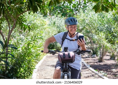 Active Smiling Senior Woman Cycling With Her Electro Bike In A Green Park Wearing Helmet. Mature Attractive Woman Stops Using Cellphone Reading Message. Retirement And Healthy Lifestyle Concept