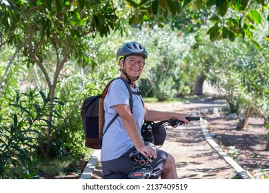 Active Smiling Senior Woman Cycling With Her Electro Bike In A Green Park Wearing Helmet Looking Back. Mature Attractive Woman Enjoying Retirement And Healthy Lifestyle. Tropical Park