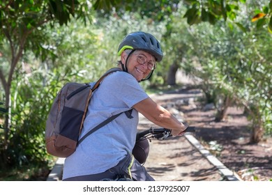 Active Smiling Senior Woman Cycling With Her Electro Bike In A Green Park Wearing Helmet Looking Back. Mature Attractive Woman Enjoying Retirement And Healthy Lifestyle. Tropical Park