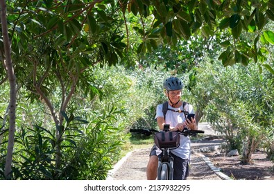 Active Smiling Senior Woman Cycling With Her Electro Bike In A Green Park Wearing Helmet. Mature Attractive Woman Stops Using Cellphone Reading Message. Retirement And Healthy Lifestyle Concept