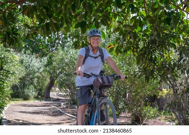 Active Smiling Senior Woman Cycling With Her Electro Bike In A Green Park Wearing Helmet. Mature Attractive Woman Enjoying Retirement And Healthy Lifestyle. Tropical Park