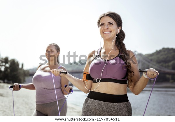 Active sport. Happy\
delighted woman using a skipping rope while standing on the beach\
with her friend