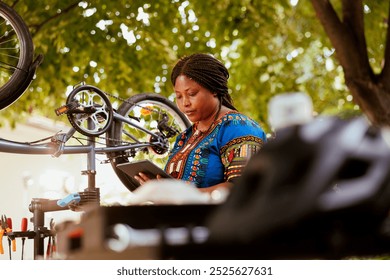 Active sports-loving black woman browsing internet on phone tablet while fixing bike wheels. Young african american individual holding digital device for bicycle repair instructions. - Powered by Shutterstock