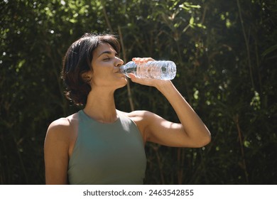 Active sporty fit young Hispanic healthy woman wearing sportswear holding bottle drinking water hydrating body after doing sport fitness training standing in green nature park outdoor, hydration. - Powered by Shutterstock