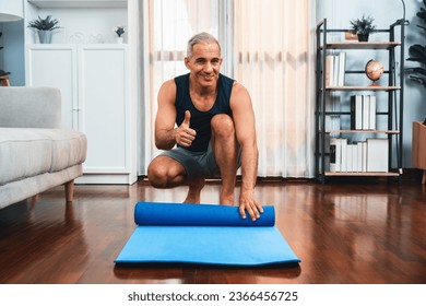 Active and sporty senior man preparing, rolling fitness exercising mat on living room floor at home. Home exercise as concept of healthy fit body lifestyle after retirement. Clout - Shutterstock ID 2366456725