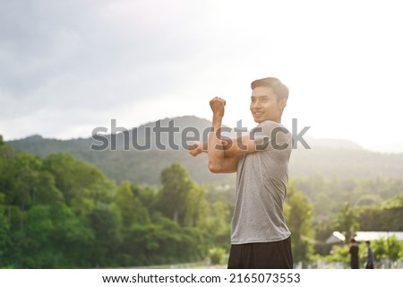 Active and sporty young asian male in sportswear stretching his arms or warming up his body before running in the green park. Sport activities concept