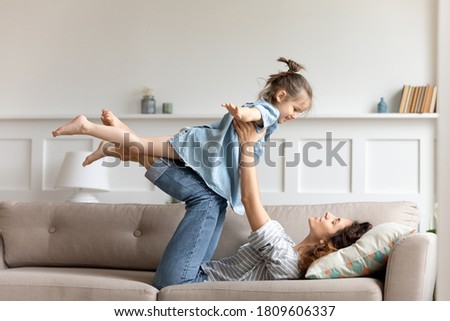 Active time with preschool child at home, easy comfort travel together with kid concept. Mother lying on couch lift up little daughter she raised hands imagines herself like plane enjoy flying in air