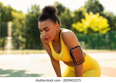 Active Training. Tired African American young woman in yellow sportswear bra and arm band with cell resting during morning run, catching breath standing outdoors at public park or sports field - Powered by Shutterstock