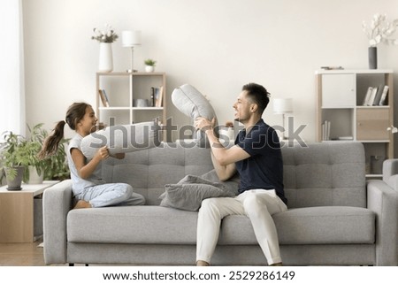 Active weekend. Naughty hyperactive little girl play on couch with grown up man father babysitter elder brother having fun together fighting with sofa cushions use orthopedic pillows for funny battle