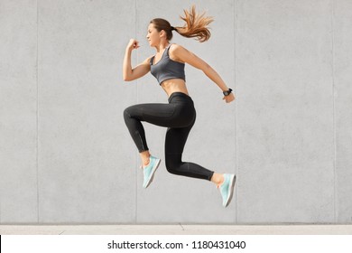 Active woman being full of energy, jumps high in air, wears sportsclothes, prepares for sport competitions, isolated over grey concrete wall. Female trainer busy with training. Gymnastics concept - Shutterstock ID 1180431040