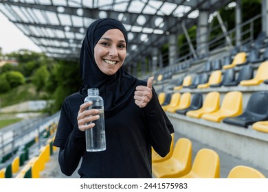 Active woman in a hijab holding a water bottle, giving a thumbs up at an outdoor stadium, showing hydration and health. - Powered by Shutterstock