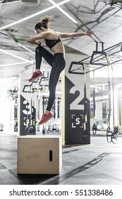 Active Woman Jumps Above The Wooden Box In The Gym On The Background Of The Hanging TRX Straps. She Wears Dark Top And Pants, Red Sneakers. Vertical.