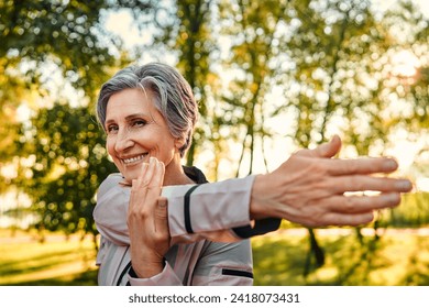 Active workout outdoors. Close up of pleasant grey haired woman in sportswear doing stretching exercises for arms at green sunny park. Caucasian lady of old ages taking care of health and body. - Powered by Shutterstock