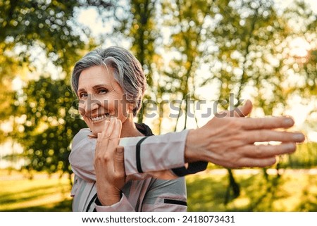 Active workout outdoors. Close up of pleasant grey haired woman in sportswear doing stretching exercises for arms at green sunny park. Caucasian lady of old ages taking care of health and body.