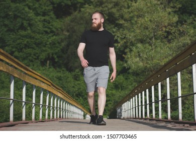 Active Young Adult Man With Beard Wearing Black Shirt And Sneakers Walking Along Wood Bridge In Natural Parkland During Cardio Workout