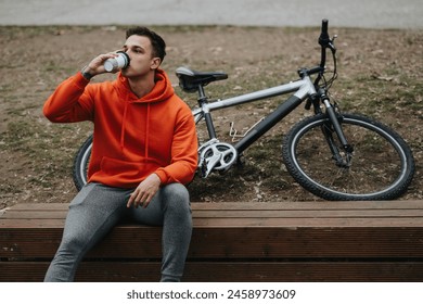 Active young adult resting on a bench in a park, drinking from a cup, with a mountain bike beside him, on an overcast day - Powered by Shutterstock