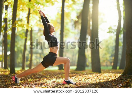 Active young female dressed in sport clothing doing stretching exercises for body on fresh air. Healthy and fit woman training regularly at local park.