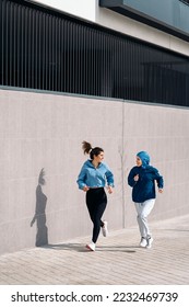 Active young female friends running in the street and smiling. One of them is wearing a hijab. - Shutterstock ID 2232469739