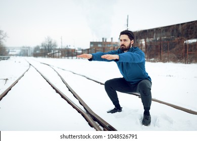 Active young man crouching and doing exercises in the public place among old railroad during the winter training outside in. Copy space.