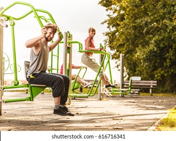 Active Young Man Exercising On Chest Press Machine And Woman On Leg Raise. Muscular Strong Guy And Girl In Training Suit Working Out At Outdoor Gym. Sport Fitness And Healthy Lifestyle Concept.